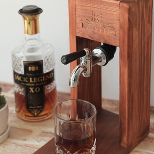 Może przedstawiać: Drewniany dozownik do alkoholu z personalizowanym napisem "KRISTIAN" z przodu. Dozownik ma metalowy kran, a do szklanki nalewany jest alkohol. W tle znajduje się butelka alkoholu Jack Legend XO.