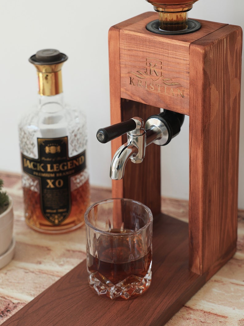 Może przedstawiać: Drewniany dozownik do alkoholu z personalizowanym napisem "KRISTIAN" z przodu. Dozownik ma metalowy kran, a do szklanki nalewany jest alkohol. W tle znajduje się butelka alkoholu Jack Legend XO.