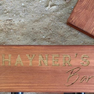 Może przedstawiać: Drewniany znak z wygrawerowanym tekstem "HAYNER'S Bar" złotymi literami. Znak jest zabarwiony na czerwono-brązowy kolor.