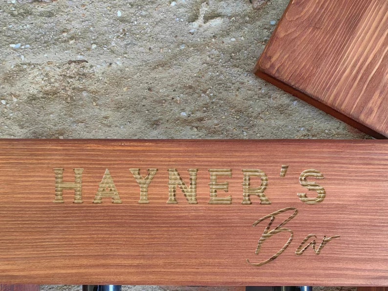 Może przedstawiać: Drewniany znak z wygrawerowanym tekstem "HAYNER'S Bar" złotymi literami. Znak jest zabarwiony na czerwono-brązowy kolor.