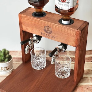 Może przedstawiać: Drewniany dozownik do alkoholu z dwoma butelkami brandy, dwoma szklankami i drewnianą podstawą. Dozownik ma dwa krany i metalową tarczę z wygrawerowanym napisem "BRANDY".