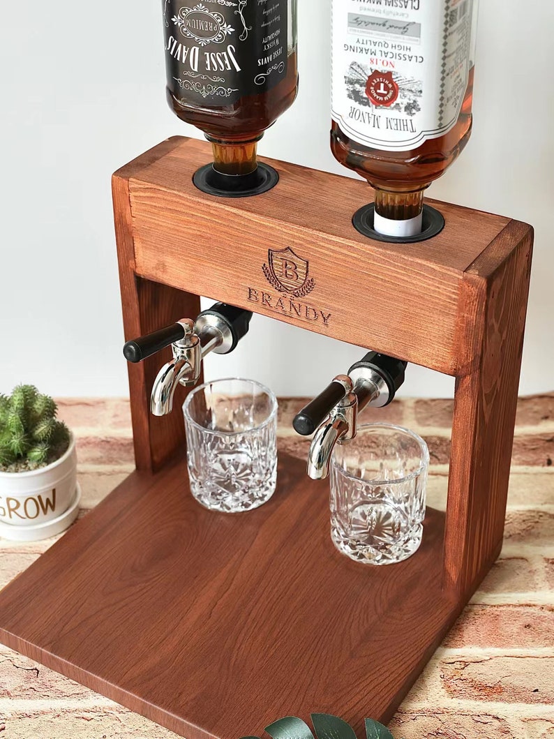 Może przedstawiać: Drewniany dozownik do alkoholu z dwoma butelkami brandy, dwoma szklankami i drewnianą podstawą. Dozownik ma dwa krany i metalową tarczę z wygrawerowanym napisem "BRANDY".