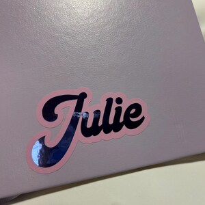Può includere: Un adesivo rosa e nero con il nome "Julie" scritto in corsivo.