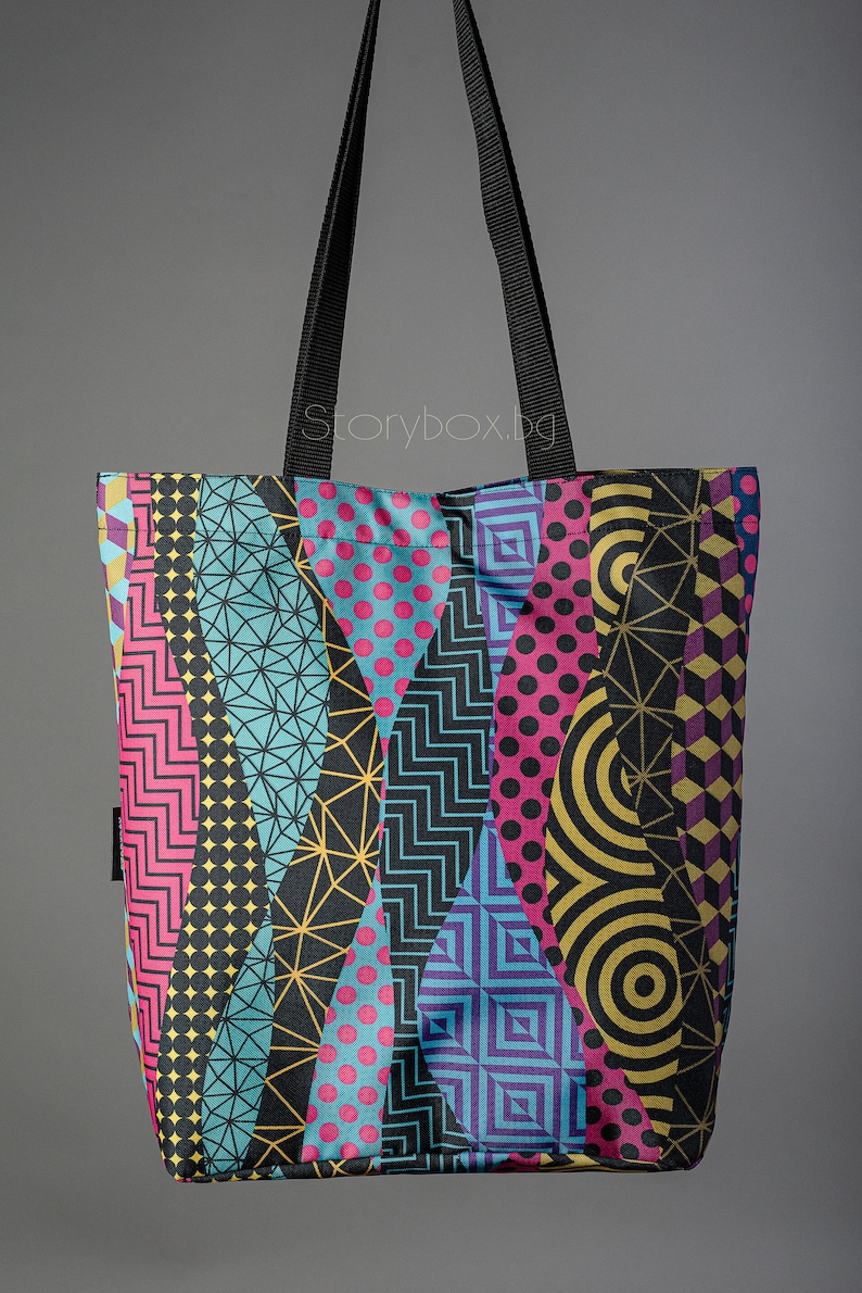 Puede incluir: Una bolsa de mano negra con un patr&oacute;n geom&eacute;trico colorido. El patr&oacute;n incluye formas rosas, azules, amarillas y moradas. La bolsa tiene correas negras.