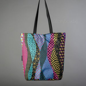 Puede incluir: Una bolsa de mano negra con un patr&oacute;n geom&eacute;trico colorido. El patr&oacute;n presenta formas geom&eacute;tricas azules, rosas, amarillas y negras.