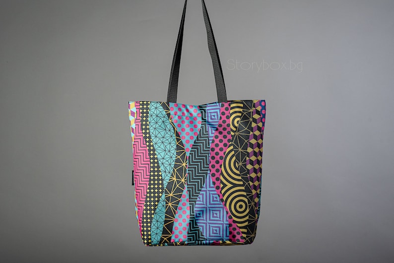 Puede incluir: Una bolsa de mano negra con un patr&oacute;n geom&eacute;trico colorido. El patr&oacute;n presenta formas geom&eacute;tricas azules, rosas, amarillas y negras.