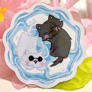 May include: A sticker featuring two cartoon cats. One cat is white with sunglasses, and the other is dark gray. They are set against a blue and white wavy background. The sticker has a scalloped edge and is placed on a pink and green surface.