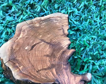 May include: A round wooden serving board made from olive wood. The board has a natural wood grain pattern and a rough, rustic texture.