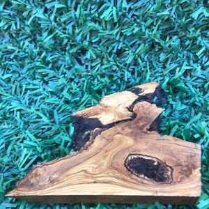 May include: A natural wood cutting board with a unique shape and grain pattern. The board has a dark brown center and lighter brown edges. It is made of olive wood.