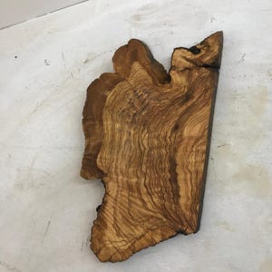 May include: A handcrafted, irregularly shaped wooden serving board. The board is made of olive wood, showcasing a rich, warm brown color with intricate grain patterns. The edges have a natural, organic shape, adding to its rustic appeal.