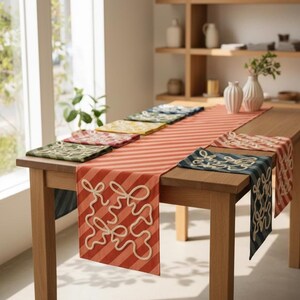 May include: A wooden table with a red and white striped table runner and matching placemats. The runner and placemats feature a white abstract design. The table is set in a well-lit room with a window and shelves in the background.