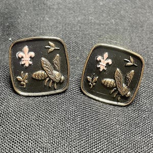 Puede incluir: Un par de pendientes cuadrados negros con detalles dorados y rosados. Cada pendiente presenta una abeja estilizada con alas, una flor de lis y hojas.