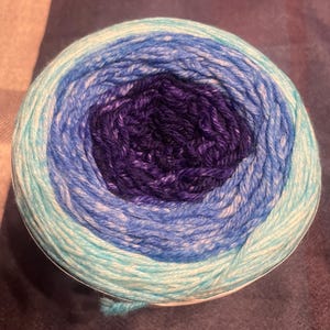 May include: A ball of yarn with a gradient of colors, including teal, light blue, dark blue, and purple. The yarn is tightly wound, showing the different color layers. Ideal for knitting and crochet projects.