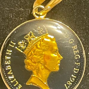 Puede incluir: Colgante dorado con diseño de moneda circular. La moneda presenta un perfil coronado con las inscripciones "ELIZABETH II" y "REG.F.D.1987". El colgante tiene un lazo dorado para sujetarlo a una cadena.