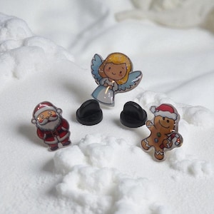 Könnte beinhalten: Drei weihnachtliche Emaille-Anstecknadeln: Ein Weihnachtsmann im roten Anzug, ein Engel mit blauen Flügeln und weißem Kleid und ein Lebkuchenmann mit Zuckerstange. Die Anstecknadeln haben schwarze Rückseiten und sind vor weißem, schneebedecktem Hintergrund platziert.