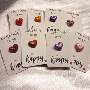 Puede incluir: Varias tarjetas con adornos en forma de corazón. Cada tarjeta tiene la frase "Little Hug to go" y "Be happy". Los corazones son de varios colores, incluyendo rojo, naranja, morado y rosa.