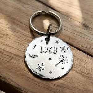 Può includere: Un medaglietta rotonda argentata per animali domestici con il nome "Lucy" inciso al centro. La medaglietta è adornata con illustrazioni fantasiose, tra cui stelle, una bacchetta magica e ali. Un portachiavi argentato è attaccato in alto.