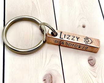 Könnte beinhalten: Ein kupferfarbener Metall-Schlüsselanhänger mit einer rechteckigen Stange, die den eingravierten Namen LIZZY, ein Pfotenabdruck-Symbol und die Zahlenfolge 074 5010872 trägt. Die Stange ist an einem silberfarbenen Schlüsselring befestigt.