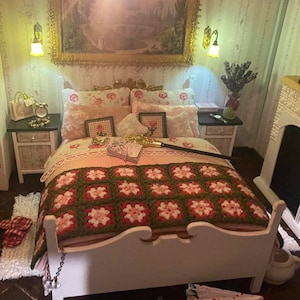 Peut inclure: Un lit blanc avec une couverture en crochet rose et verte. Le lit a une tête de lit et un pied de lit blancs avec un motif décoratif. Il y a plusieurs oreillers sur le lit, dont deux avec des motifs floraux et deux avec des couleurs unies. Le lit est dans une pièce avec une cheminée et une table de chevet.
