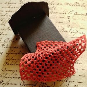 Peut inclure: Un lit miniature en carton avec une couverture en crochet rouge. Le lit est posé sur un morceau de papier avec du texte manuscrit.