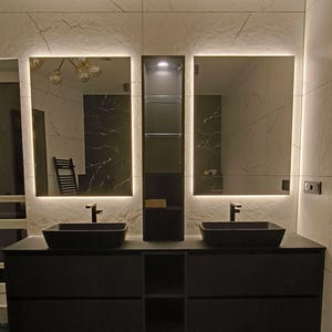 Könnte beinhalten: Zwei schwarze Badezimmerwaschbecken mit verchromten Armaturen, montiert auf einem schwarzen Waschtisch mit offenen Regalen. Die Waschbecken befinden sich vor drei Spiegeln mit weißen LED-Leuchten um die Ränder. Die Wände sind aus weißem Marmor.