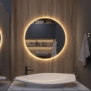 Può includere: Uno specchio da bagno rotondo con una luce LED bianca calda attorno al bordo. Lo specchio è montato su una parete in legno e riflette uno scaffale con articoli da toeletta e asciugamani.