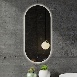 Puede incluir: Un espejo ovalado moderno con un fino borde iluminado y una pequeña lámpara redonda blanca que cuelga de la parte superior. El espejo está montado en una pared de hormigón gris.