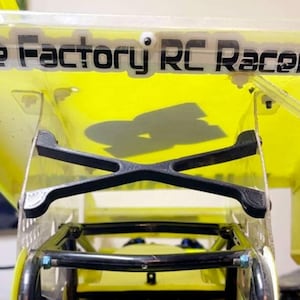 May include: A close-up of a yellow RC car wing with the text "The Factory RC Raceway" in black. The wing is supported by a black cross-shaped structure. The car's frame is visible below.