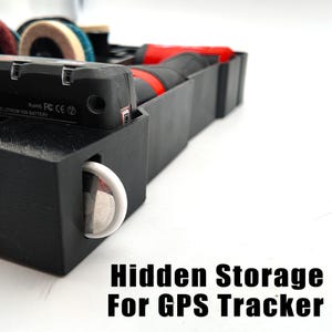 May include: Black plastic hidden storage container for a GPS tracker. The container has a compartment for the tracker and a slot for a white circular object. The text "Hidden Storage For GPS Tracker" is at the bottom.