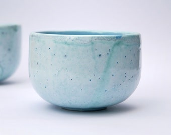 May include: A light blue ceramic bowl with a speckled glaze. The bowl is round and has a slightly flared rim.