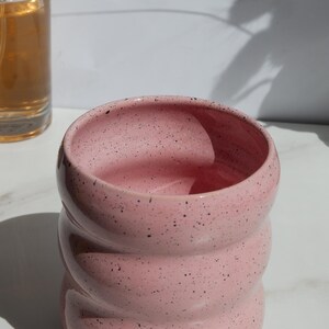 May include: A pink ceramic cup with a speckled finish and a distinct rippled, wavy texture along its exterior. The vessel features a wide, open circular rim and a rounded body, showcasing a smooth, glossy glaze with small black flecks throughout.