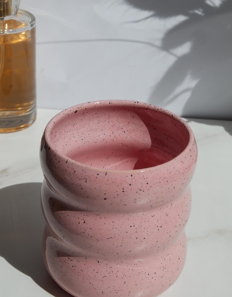May include: A pink ceramic cup with a speckled finish and a distinct rippled, wavy texture along its exterior. The vessel features a wide, open circular rim and a rounded body, showcasing a smooth, glossy glaze with small black flecks throughout.