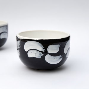 May include: Two black ceramic bowls with white abstract brushstroke designs. The bowls are round and have a slightly flared lip.