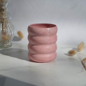 Pode incluir: Uma caneca de cerâmica rosa com um design texturizado e canelado e acabamento salpicado. A caneca é cilíndrica com uma abertura larga. O fundo inclui um vaso de vidro e flores secas, tudo sobre uma superfície de mármore branco.