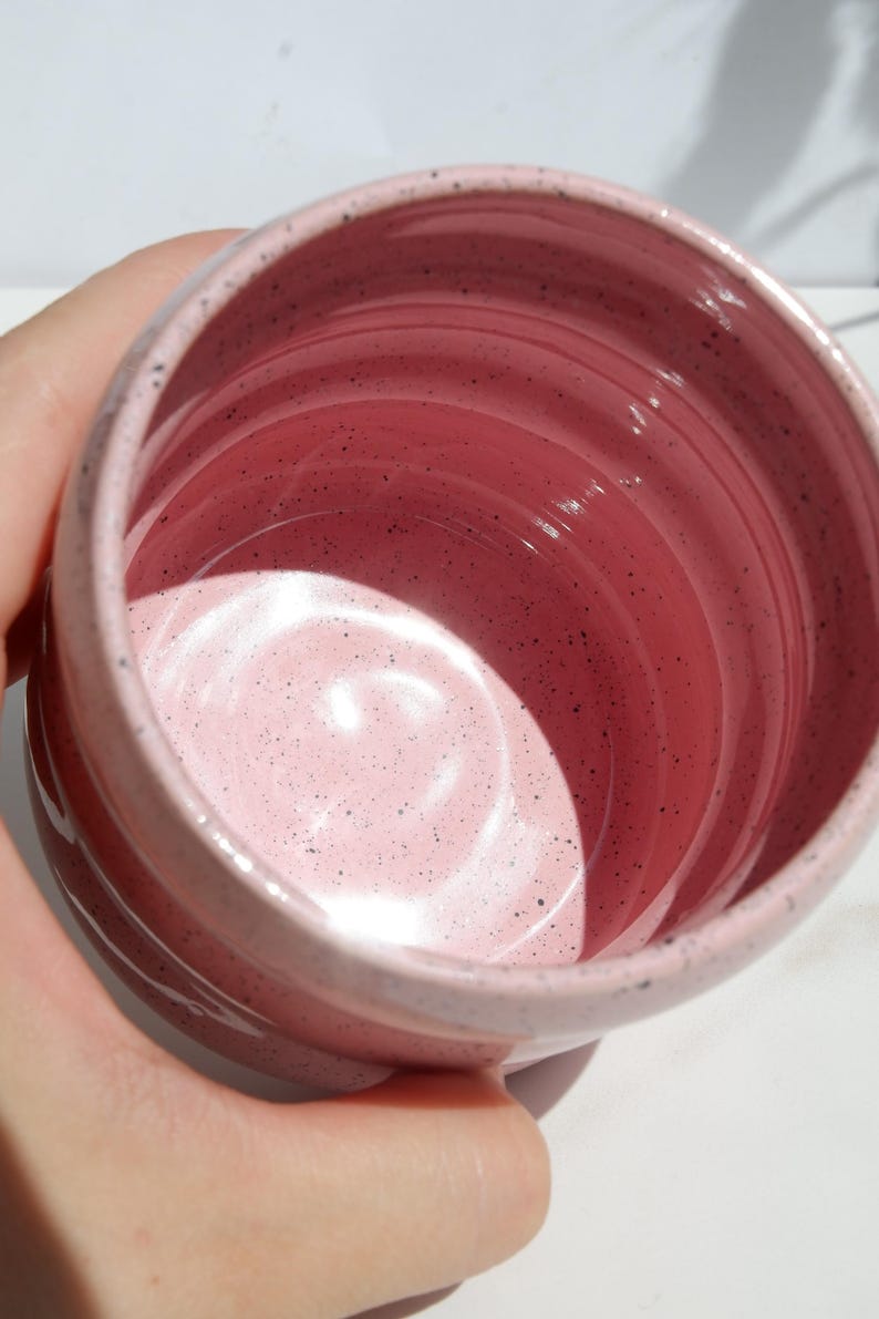 May include: A cylindrical ceramic mug with a glossy pink glaze and dark speckles throughout the interior and exterior. The vessel features a smooth, rounded rim and subtle horizontal ridges along the inner walls, creating a simple, functional design.