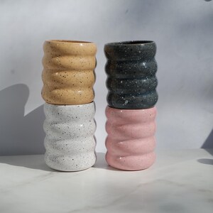 May include: Four cylindrical ceramic cups with a wavy, ribbed exterior texture. The cups are arranged in two stacks of two. The colors include a speckled tan, a speckled white, a speckled dark gray, and a solid light pink finish.