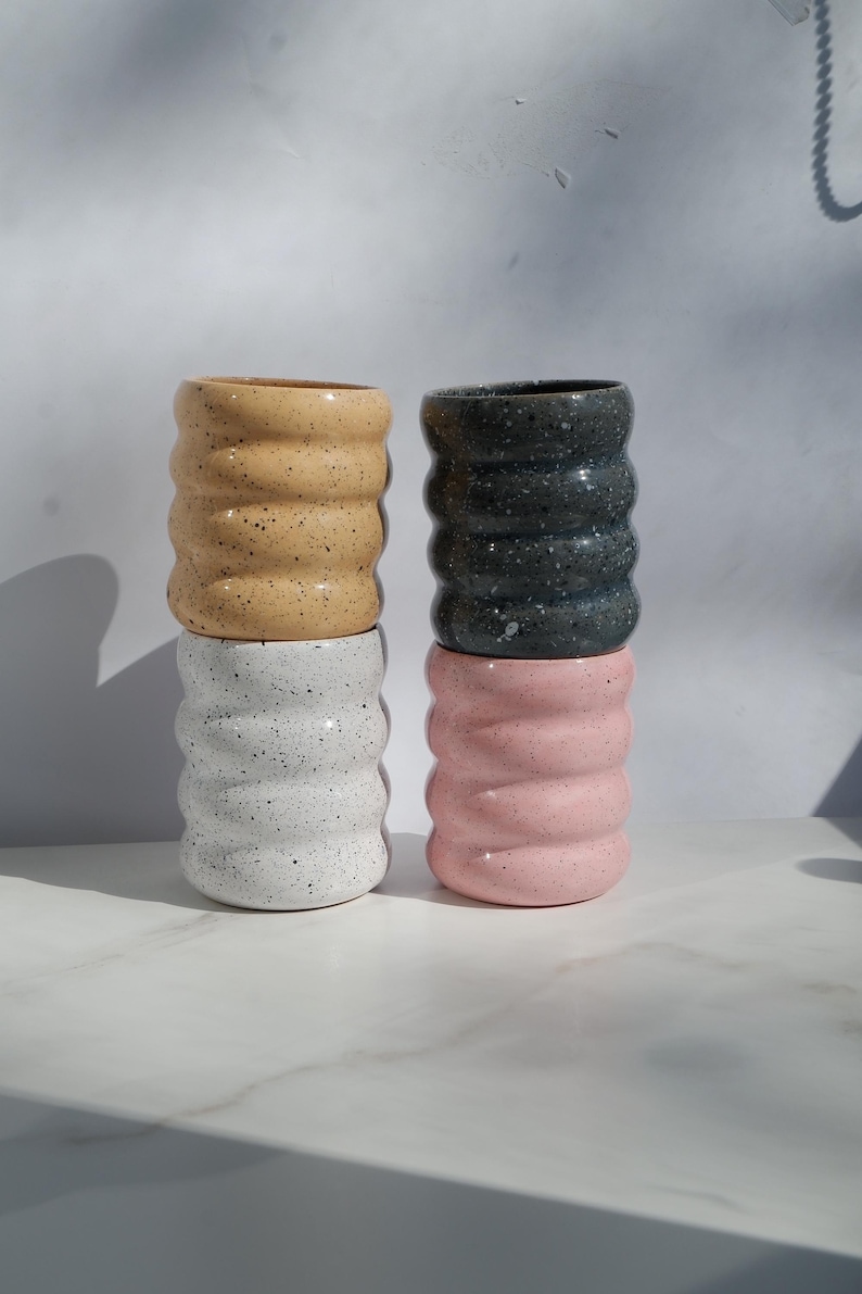 May include: Four cylindrical ceramic cups with a wavy, ribbed exterior texture. The cups are arranged in two stacks of two. The colors include a speckled tan, a speckled white, a speckled dark gray, and a solid light pink finish.