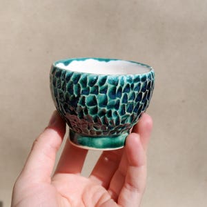 Könnte beinhalten: Eine kleine, handgefertigte Keramikschale mit einer strukturierten, mosaikartigen Außenseite in Türkis und Dunkelblau. Der Innenraum ist weiß. Die Schale wird in einer Hand gehalten und zeigt ihr einzigartiges Design und ihre Handwerkskunst.