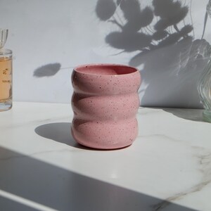 May include: A cylindrical ceramic cup with a soft pink glaze and dark speckles throughout. The exterior features three distinct horizontal rounded ridges that create a wavy, undulating profile from the base to the wide, open rim.