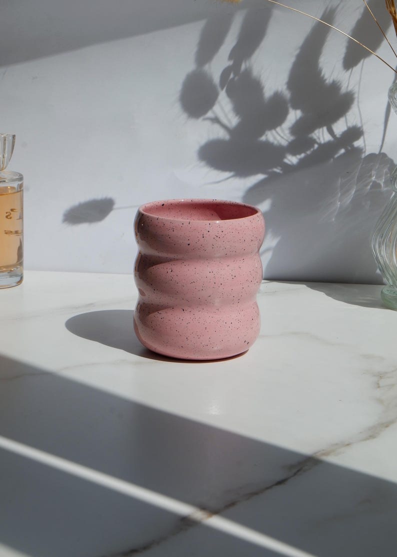 May include: A cylindrical ceramic cup with a soft pink glaze and dark speckles throughout. The exterior features three distinct horizontal rounded ridges that create a wavy, undulating profile from the base to the wide, open rim.