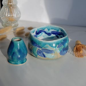 Könnte beinhalten: Eine Keramik-Matcha-Schale mit blauem und türkisfarbenem Wellenmuster, eine passende teal-farbene Teedose und ein Bambusbesen. Die Schale hat einen hellbeigen Boden und ist mit blauen Meereskreaturen verziert.