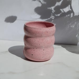 May include: A cylindrical ceramic cup with a light pink glaze and dark speckles throughout. The vessel features three distinct horizontal ridges or indentations along its body, creating a wavy, rounded profile with an open top and a flat base.