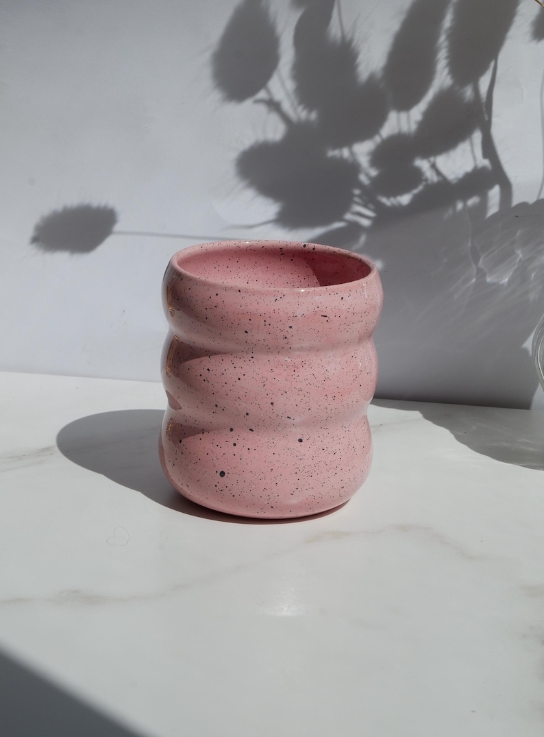May include: A cylindrical ceramic cup with a light pink glaze and dark speckles throughout. The vessel features three distinct horizontal ridges or indentations along its body, creating a wavy, rounded profile with an open top and a flat base.