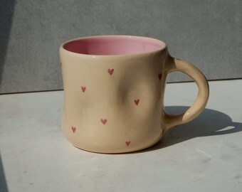 May include: A beige ceramic mug with a pink interior and small red heart designs. The mug has a rounded shape and a curved handle. The mug is sitting on a white surface with a gray background.
