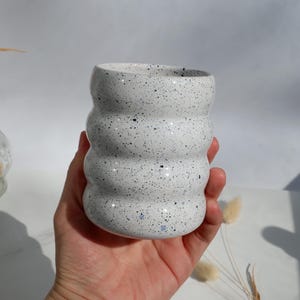 Op de afbeelding: Een witte keramische vaas met een gespikkeld patroon van zwarte en blauwe stippen. De vaas heeft een uniek, afgerond, gestapeld ontwerp. De vaas wordt in een hand gehouden.