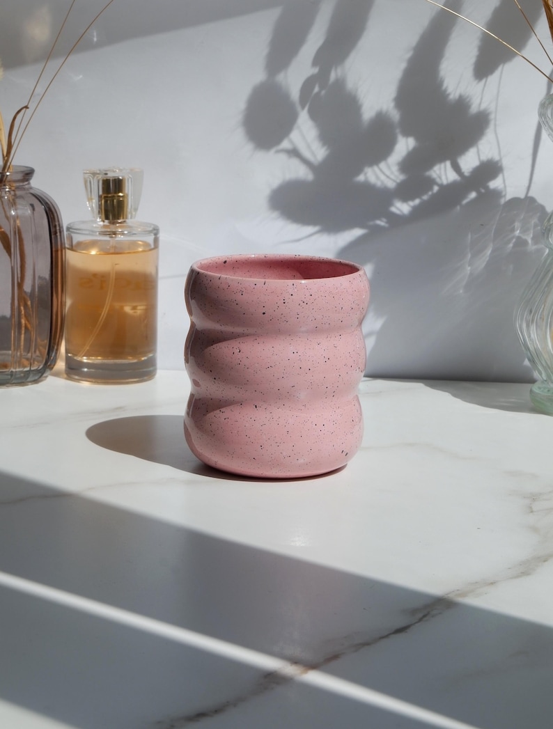 May include: A cylindrical ceramic cup with a light pink glaze and dark speckles throughout. The vessel features three distinct horizontal rounded ridges or ripples along its exterior surface, creating a wavy, textured profile from top to bottom.