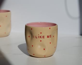 May include: A beige ceramic mug with a pink interior and red heart accents. The mug has the text "I LIKE ME" in red. Another mug is slightly visible in the background. The mug is on a white surface.