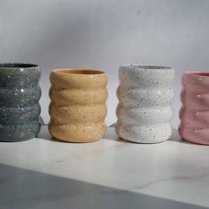 May include: Four cylindrical ceramic cups with a wavy, ribbed exterior texture. Each cup features a speckled finish in distinct colors: dark gray, light tan, white, and soft pink. The cups are arranged in a horizontal row on a flat surface.