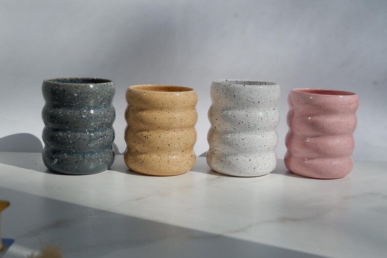 May include: Four cylindrical ceramic cups with a wavy, ribbed exterior texture. Each cup features a speckled finish in distinct colors: dark gray, light tan, white, and soft pink. The cups are arranged in a horizontal row on a flat surface.