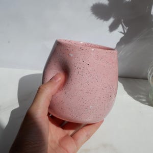 May include: A light pink ceramic cup with a speckled finish featuring small black and white dots. The cup has a rounded, bulbous shape with a distinct indentation on one side, creating a curved, ergonomic form for holding the vessel comfortably.